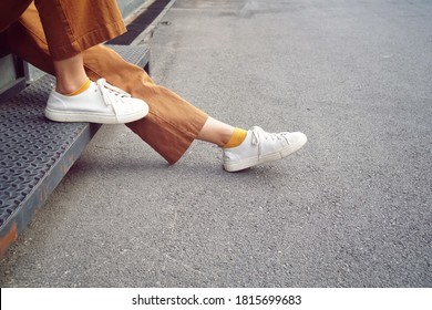 Low Section Of Body Woman Sitting On Metal Staircase And Feet Placed On Road, Dress Up With Light Brown Trouser, Yellow Mustard Sock And White Sneaker, With Copy Space, Street Style Fashion