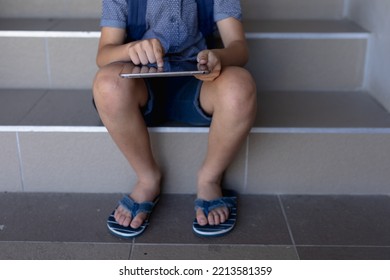 Low Section Of A Caucasian Schoolboy Wearing Flip-flops And Shorts Sitting On Steps At Elementary School Using A Tablet Computer