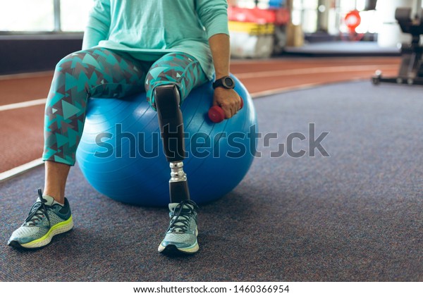 Low section of disabled active senior Caucasian woman\
with leg amputee exercising with dumbbell while sitting on exercise\
ball in fitness studio. Strong active senior female amputee\
training and