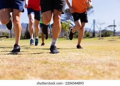 Low section front view of the legs of a multi-ethnic group of male runners training at a sports field, running together on a grass track in the sun