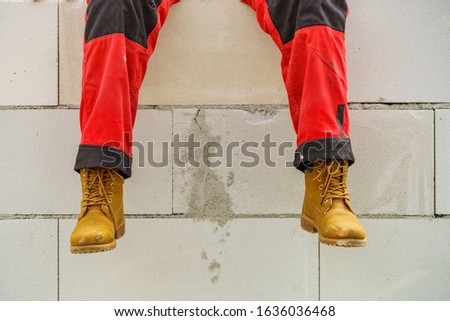 Low section legs of construction worker wearing workwear trousers and brown leather work boots relaxing on airbrick wall.