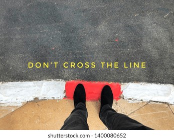 Low Section Of Man Standing On A Red  Line With The Text : Don't Cross The Line Written On Asphalt