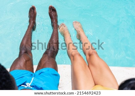 Low section of multiracial young couple dangling legs in swimming pool. unaltered, togetherness, lifestyle, leisure, bonding, weekend activities.