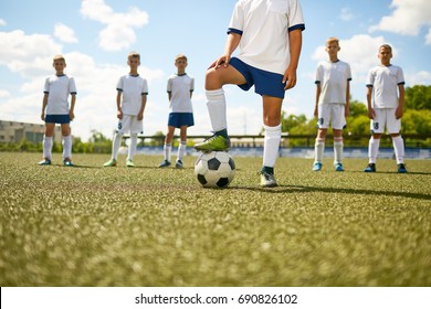 Low Section Portrait Of Football Captain Standing In Center Of Field Stepping On Ball With The Rest Of Team In Background