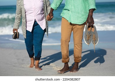 Low Section Of A Senior African American Couple Walking Barefoot On The Beach With Blue Sea In The Background, Holding Hands And Their Shoes 