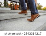 Low section of senior male entrepreneur in brown leather shoes moving up on staircase in modern city