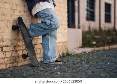 Low section of a skater leaning against a brick wall with their skateboard propped up beside them, showcasing urban street style and skateboarding culture - Powered by Shutterstock