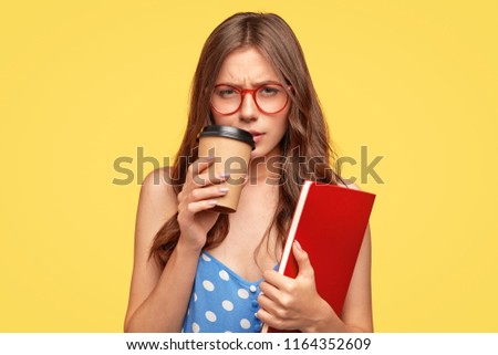 Low spirited young female student feels sad after recieving bad mark for exam, being tired, drinks hot coffee, holds scientific literature, stands against yellow background. Youth, studying concept