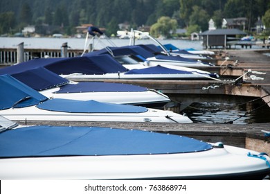 Low telephoto view on the bows and windshields of luxury water skiing motor boats protected by blue canvas covers and docked in row, tied along a wood pier with lake and shore in background