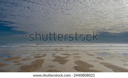 The low tide in the ocean. Rivulets of water form a bizarre pattern on the sand. Turquoise waves roll towards the shore, foaming and spreading along the beach. Beautiful clouds in the blue sky. 