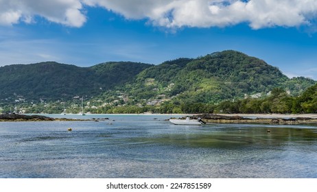 Low tide on a tropical island. The white boat is moored in shallow water. The stones of the exposed seabed are visible. Green hills against a blue sky of clouds. Seychelles. Mahe. Beau Vallon - Shutterstock ID 2247851589