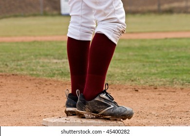 Low View Of Baseball Player Standing On Base With Muddy Cleats, Dark Red Socks And White Pants