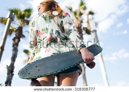 Low view of a hipster girl holding long board with copy space area for your brand while standing against sky and palm trees resting after riding, young woman with skateboard relaxing after skating