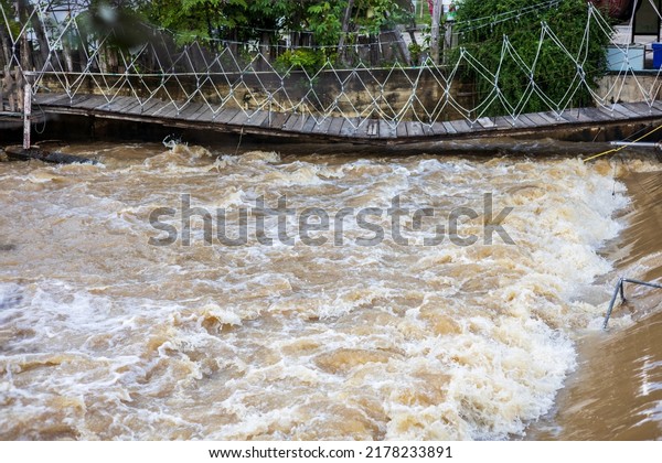 A low view of the river flowing violently past\
a broken wooden bridge near the bank of an old wooden fence inside\
a temple in rural Thailand.