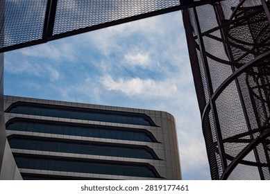 A low-angle abstract architectural composition captures a modern high-rise office building framed by structural steel mesh elements under a bright blue sky filled with wispy white clouds. The sleek concrete exterior and dark tinted horizontal windows of the office tower create a sharp contrast with the copper-toned curved metal lattice in the foreground. Strong geometric lines and open sky give the image a spacious, forward-thinking feel. This creative urban view is ideal for corporate real estate brochures, architectural showcases, construction firm branding, commercial business presentations, and modern city planning stock photography collections.