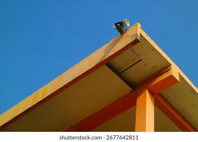 A low-angle abstract shot of the corner of a public building's roof with a loudspeaker attached, set against a vast and clear blue sky. The photo captures a unique perspective of urban architecture