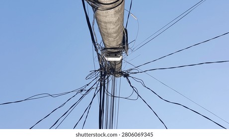 A low-angle abstract view of a concrete utility pole with a chaotic, tangled mess of overhead power lines and telecommunication cables against a clear blue sky. A vivid representation of urban infrastructure challenges, network complexity, and messy cable management.