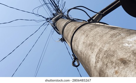 A low-angle abstract view of a concrete utility pole with a chaotic, tangled mess of overhead power lines and telecommunication cables against a clear blue sky. A vivid representation of urban infrastructure challenges, network complexity, and messy cable management.