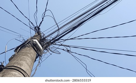 A low-angle abstract view of a concrete utility pole with a chaotic, tangled mess of overhead power lines and telecommunication cables against a clear blue sky. A vivid representation of urban infrastructure challenges, network complexity, and messy cable management.
