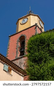 A low-angle close view shows the red and ochre bell tower with visible clocks, a bell opening, dense green ivy, a tiled roof and a clear blue sky in bright sunlight. Saint-Tropez, Var, Provence-Alpes-Cote d Azur, France