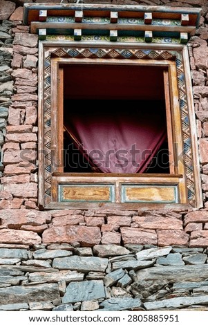A low-angle close-up capturing an open wooden window frame built into a rugged, dry-stacked masonry wall made of coarse pink and brown fieldstones. The polished natural wood frame is bordered by a delicately painted geometric pattern in shades of blue, yellow, and white. Above the opening, an ornate overhanging eave displays stacked timber corbels with colorful painted accents. Inside the window, a triangular fold of deep magenta fabric is visible, adding a vibrant pop of color against the earthy, historical stone texture.
