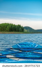 A low-angle closeup shows the patterned blue deck of a stand up paddleboard floating on calm water looking toward distant green trees and hills.