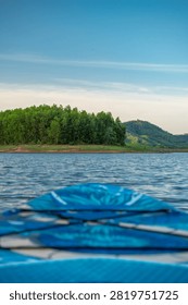 A low-angle closeup shows the patterned blue deck of a stand up paddleboard floating on calm water looking toward distant green trees and hills.
