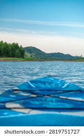 A low-angle closeup shows the patterned blue deck of a stand up paddleboard floating on calm water looking toward distant green trees and hills.