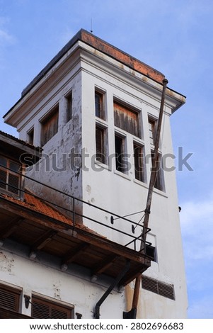 Low-angle close-up of weathered white colonial tower with peeling plaster, rusty roof and wooden louvered windows against blue sky in Jakarta
