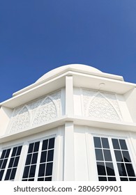 Low-angle corner view of a modern white building with a dome, Islamic geometric relief patterns, and grid windows against a vivid blue sky.