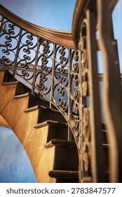 A low-angle, diagonal perspective shot of a classic curved spiral staircase. The composition highlights textured golden stone steps rising alongside an intricate wrought iron balustrade featuring elegant scroll patterns. A polished wooden handrail sweeps gracefully through the upper frame, contrasting against a clear blue sky background. The vertical structure and rich textures evoke timeless architectural design, luxury residential interiors, and traditional craftsmanship.