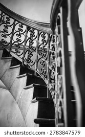 A low-angle, diagonal perspective shot of a classic curved spiral staircase. The composition highlights textured golden stone steps rising alongside an intricate wrought iron balustrade featuring elegant scroll patterns. A polished wooden handrail sweeps gracefully through the upper frame, contrasting against a clear blue sky background. The vertical structure and rich textures evoke timeless architectural design, luxury residential interiors, and traditional craftsmanship.