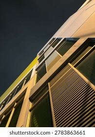 A low-angle, dramatic perspective of a modern building facade stretching upward against a dark night sky. The architectural shot highlights geometric lines, multiple glass windows, a section with horizontal louvers  and warm exterior lighting casting a glow on the structure's surfaces.