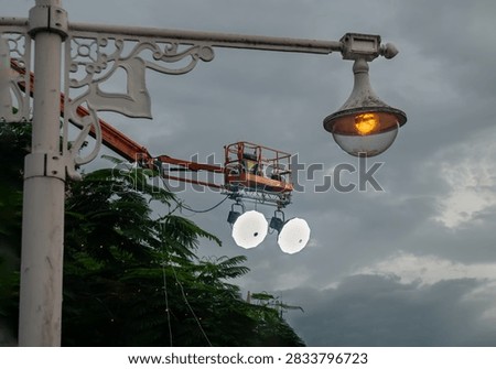 A low-angle evening shot capturing a white decorative streetlamp post adorned with classical scrollwork, emitting a soft, golden light. In the background, an industrial cherry picker crane extends an orange boom arm into the frame. Suspended beneath the worker basket are two powerful, white-hot halogen utility lamps that cast a brilliant glare against the dark, dramatic clouds. Lush green tree foliage borders the lower-left corner, anchoring the blend of vintage aesthetics and modern urban maintenance.
