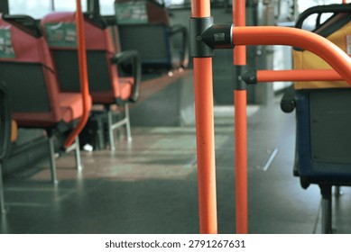 A low-angle, eye-level interior view of an empty public bus, focusing on the bright orange safety handrails and poles in the foreground. The background features rows of blurred, empty passenger seats with red and blue fabric. The lighting suggests daytime travel, conveying a sense of quiet solitude, urban commuting, transit, and public transport infrastructure. This image is suitable for themes related to travel, urban planning, mobility, and social distancing.
