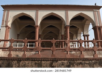 A low-angle, front-facing view captures a historic pavilion featuring cream-colored walls and prominent pointed arches supported by slender red sandstone columns. An decorative sandstone balustrade runs along the elevated terrace in the foreground above a weathered base. Clear, soft daylight overhead illuminates the geometric layout and traditional architectural details.