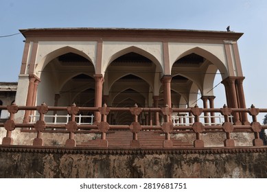 A low-angle, front-facing view captures a historic pavilion featuring cream-colored walls and prominent pointed arches supported by slender red sandstone columns. An decorative sandstone balustrade runs along the elevated terrace in the foreground above a weathered base. Clear, soft daylight overhead illuminates the geometric layout and traditional architectural details.