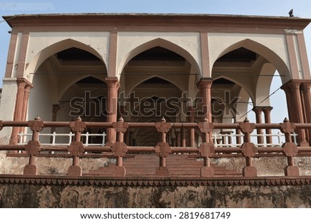 A low-angle, front-facing view captures a historic pavilion featuring cream-colored walls and prominent pointed arches supported by slender red sandstone columns. An decorative sandstone balustrade runs along the elevated terrace in the foreground above a weathered base. Clear, soft daylight overhead illuminates the geometric layout and traditional architectural details.