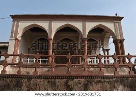 A low-angle, front-facing view captures a historic pavilion featuring cream-colored walls and prominent pointed arches supported by slender red sandstone columns. An decorative sandstone balustrade runs along the elevated terrace in the foreground above a weathered base. Clear, soft daylight overhead illuminates the geometric layout and traditional architectural details.
