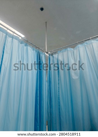 A low-angle, full shot captures a light blue privacy curtain dividing medical cubicles or examination beds inside a clinic or hospital room. The fabric is suspended by metallic rings from an L-shaped ceiling track system, which is secured and stabilized by a central vertical metal rod or chain attached to the white ceiling. The curtains are drawn closed, completely blocking the view into the patient areas. A glowing fluorescent tube light fixture is visible on the upper left side, casting a bright illumination over the scene. The overall image has a slightly soft focus, grainy texture, and subtle motion blur.