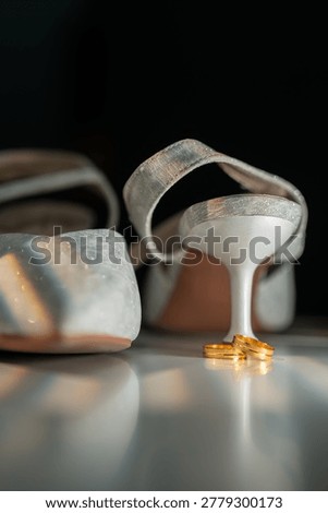 A low-angle macro shot of two gold wedding bands resting on a reflective white surface at the base of a white bridal kitten heel, with a dark black background and prismatic light flares.