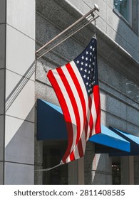 A low-angle, medium close-up shot capturing a single United States flag hanging from a metal flag pole mounted on the classic exterior of a building in the Motomachi shopping district of Yokohama, Japan. The stars and stripes of the American flag drape elegantly down, caught in a slight breeze. Behind it, the building's facade is constructed from polished light-colored stone tiles and gray granite columns, with deep blue fabric window awnings adding a rich splash of color. Bright, direct daytime sunlight casts sharp, architectural shadows across the textured stone wall, emphasizing a classic, clean aesthetic.