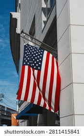 A low-angle, medium close-up shot capturing a single United States flag hanging from a metal flag pole mounted on the modern, classic exterior wall of a building in the Motomachi shopping district of Yokohama, Japan. The stars and stripes of the American flag drape down gracefully, contrasting against the clean, light-colored stone panels of the building facade. Below the flag, a deep blue fabric window awning and a small orange street banner are partially visible. In the background, utility lines, a street lamp, and a blue sky with soft, wispy clouds complete the urban scene on a bright day.