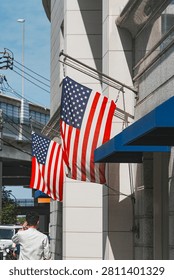 A low-angle, medium shot capturing two United States flags hanging from metal poles mounted on the exterior wall of a classic building in the Motomachi shopping district of Yokohama, Japan. The stars and stripes of the American flags drape neatly next to deep blue fabric window awnings. The building’s facade is made of light-colored stone tiles and gray granite columns. In the lower left, the back of a pedestrian in a light-colored shirt is visible as they walk down the street, while a concrete elevated road structure, utility wires, a street lamp, and a blue sky with light clouds fill the background.