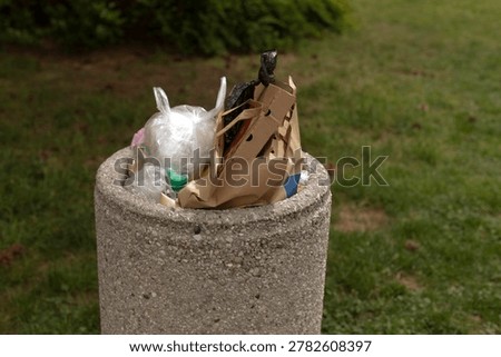 Lowangle overflowing bin with packaging and cup intimate perspective, sunlight and shadow play, textured concrete foreground and muted residential backdrop,