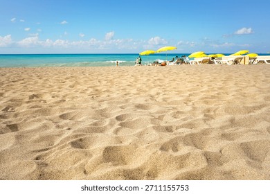 Low-angle panoramic view of row of yellow beach umbrellas and sunbeds on sandy resort beach. Vast nature scene with textured sand foreground, turquoise sea, bright blue sky. Summer holiday concept.