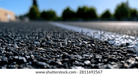 A low-angle perspective of an asphalt road, highlighting the texture of the surface and a white dividing line, with blurred trees and sky in the background.