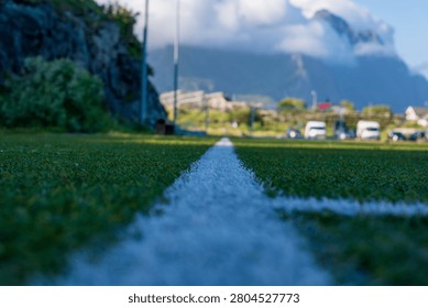 Low-angle perspective of athletic field with white boundary line, dramatic mountain backdrop and parked cars. Lofoten landscape setting