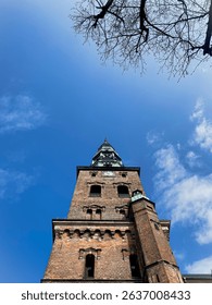 A low-angle perspective highlights the weathered red brick texture and green copper details of the Nikolaj Kunsthal church tower, evoking a grand and timeless architectural presence.
