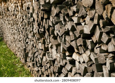 Low-angle perspective of a long wall of stacked split firewood receding into the distance. A striking, dynamic shot of a meticulously stacked wall of chopped firewood, shot from a sharp side angle. The repeating pattern of the split logs creates a powerful visual rhythm and a deep sense of perspective, as the woodpile stretches far into the background. A sliver of bright green grass at the base provides a vibrant contrast to the earthy tones of the timber. Excellent for backgrounds, abstract compositions, and rustic concept design.